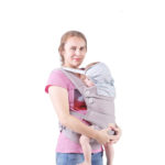 Porte bébé gris avec protection de tête en coton. Sa maman est blonde avec un t-shirt rose, sourit en maintenant son bébé par le bas du dos à l'aide de ses deux mains.