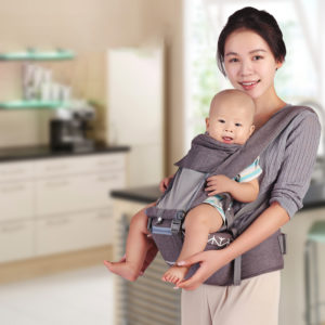 Porte bébé gris avec bébé qui sourit dos à la maman qui sourit et qui tient le pied de son bébé. En fond d'image, on devine des meubles de rangement d'un salon.
