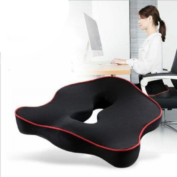 Coussin ergonomique noir avec des contours rouges. Derrière se trouve une femme brune avec un chemisier blanc à son bureau. Elle est assise sur une chaise blanche et travaille sur son ordinateur blanc.