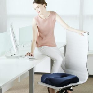 Une femme est assise sur son bureau blanc avec un ordinateur blanc posé dessus. Elle se tient à une chaise de bureau blanche avec un cousin ergonomique bleu dessus. La femme porte un pantalon blanc avec un t-shirt rose. Elle est brune et est coiffée avec un chignon.