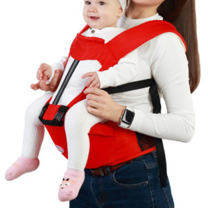 Porte bébé rouge à sangles noires avec bébé fille qui sourit à dos à sa maman. Le bébé a des petits chaussons roses.