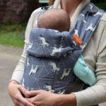 Porte bébé bleu à motif cheval blanc. bébé contre sa maman. La tête du bébé dépasse du porte bébé.