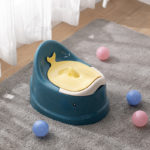 Pot pour bébé ergonomique vert foncé et couvercle jaune clair en forme de baleine. Tapis gris, boules de jeux bleu et rose au sol. Rideau voilage blanc.