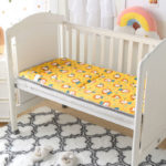 Matelas ergonomique jaune à tête de lion pour lit de bébé. Installé dans un lit blanc à barreaux. Arc en ciel en peluche au dessus et tapis à motif blanc et noir en dessous