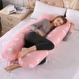 Oreiller de grossesse rose en forme de U sur lit aux draps blancs. Et femme qui dort sur le côté en legging noir qui tient le haut du coussin avec sa main gauche.
