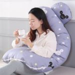 Coussin de grossesse gris plié avec femme assise dedans en train de boire un café.