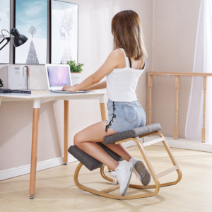 Femme de dos avec un débardeur blanc et un short en jean assise sur un siège ergonomique en bois. Elle est assise à un bureau blanc. Sur le bureau se trouve un ordinateur, une lampe un une plante. Il y a deux poster au mur.