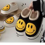 Au premier plan, main tenant des chaussons noirs avec un smiley jaune. En fond, une paire de chaussons blancs avec smiley posée sur une étagère.