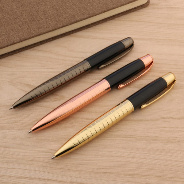 3 stylos sont posés sur une table en bois. Un est marron, l'autre rose et le dernier or. Un classeur se trouve à côté.