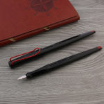 Deux stylos noir et rouge sont posés sur une table en bois. Un des stylos a un bouchon l'autre non. Un classeur en cuir marron se trouve à côté.