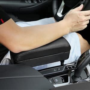On voit un accoudoir d'appoint installé entre l'accoudoir d'une voiture et le siège conducteur. Il est noir, il est ergonomique et le bras du conducteur est posé dessus.