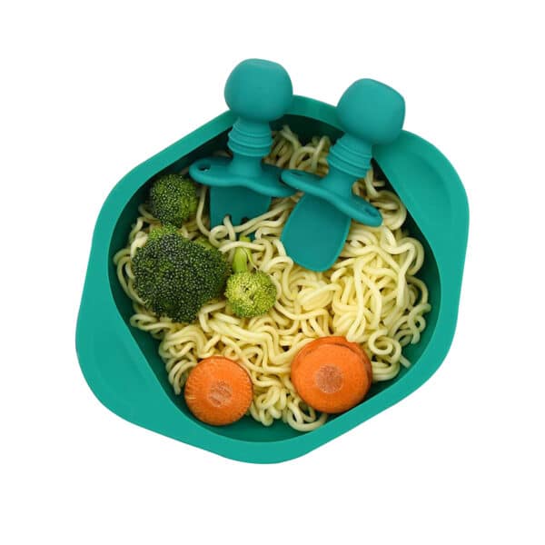 Assiette écuelle en silicone vert émeraude, remplie de spaghettis, avec deux rondelles de carottes et deux morceaux de brocoli. Et posé en haut à droite dans l'assiette une mini cuillère et une mini fourchette vert émeraude, en silicone avec un manche en forme de boule. Le tout sur un fond blanc vu de haut.