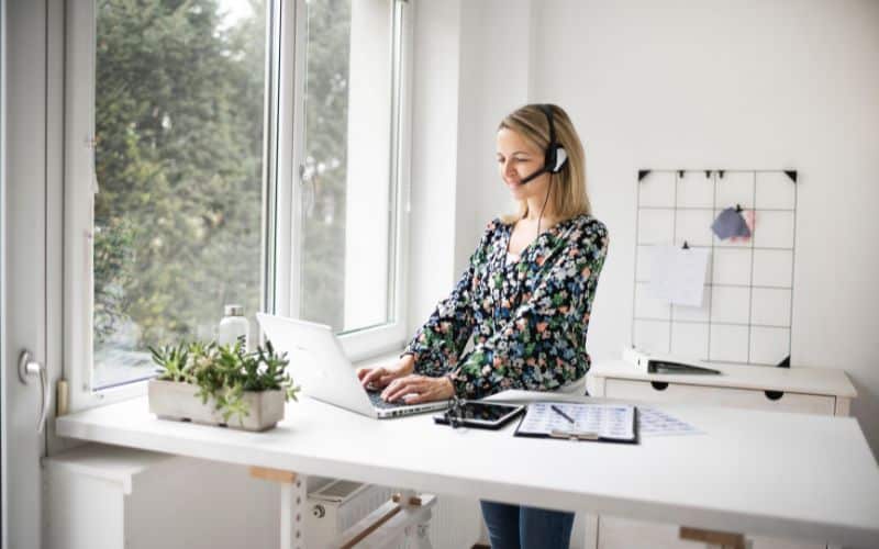 Femme debout devant son bureau avec un ordinateur, une oreillette micro et d'autres objets ergonomiques.