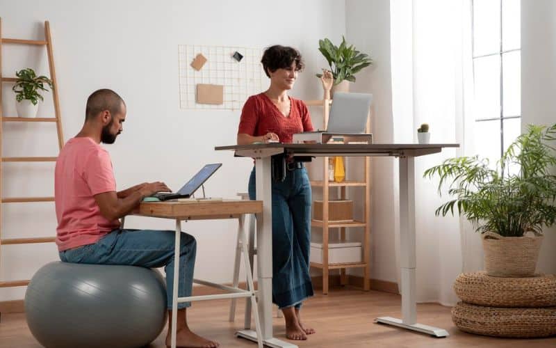 Pièce avec deux bureaux, un haut avec une femme qui est sur un ordinateur debout, et un plus bas où se trouve un homme sur un ballon qui utilise un ordinateur.