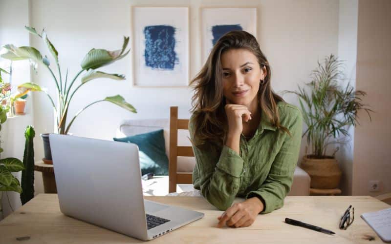 Femme assise à une table avec son ordinateur, des lunettes, un cahier et un style sont posés à côté de son bras sur la table. Derrière elle, on voit son coin salon.