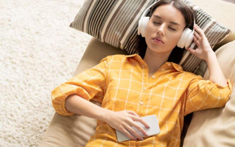 Comment les objets ergonomiques favorisent la relaxation Jeune femme allongée, la tête sur un oreiller avec un casque de musique sur les oreilles, les yeux fermés et un téléphone dans une main.