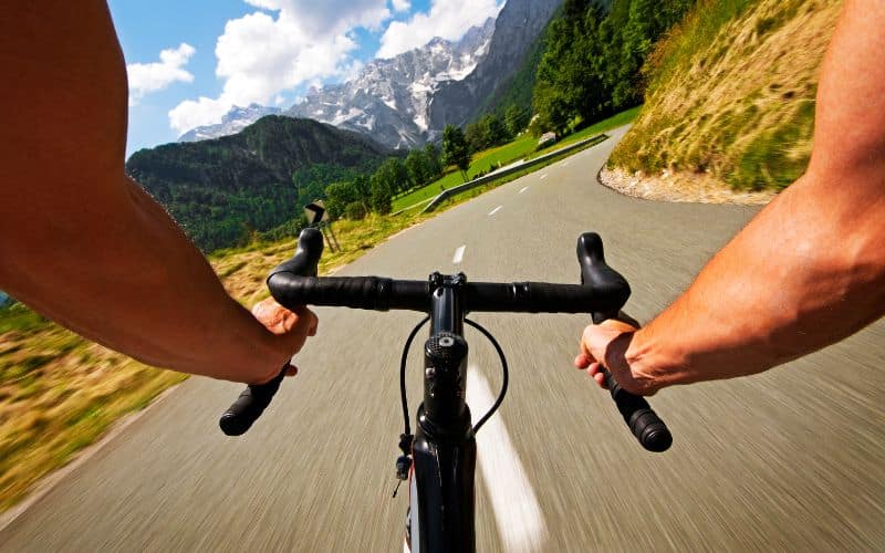 Deux bras qui tiennent un guidon de vélo ergonomique, avec une vue magnifique sur les montagnes et la route que le cycliste descend.