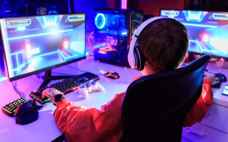 Jeune garçon assit sur une chaise gaming, face à deux écrans, avec un clavier et une souris ergonomique, une manette de console, et un casque audio sur les oreilles, il est dos à nous.