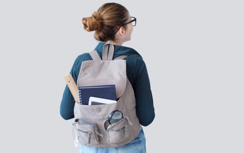 Jeune fille de dos, portant un sac à dos gris avec des affaires scolaire qui dépassent d'une poche avant.