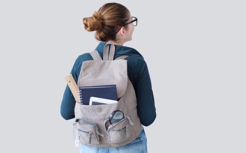 Les bienfaits d'un sac à dos ergonomique sur la posture Jeune fille de dos, portant un sac à dos gris avec des affaires scolaire qui dépassent d'une poche avant.
