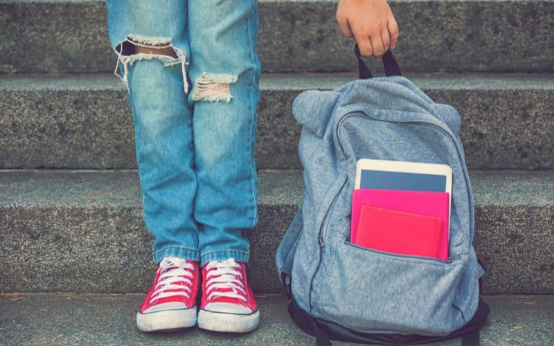 La différence entre un sac à dos classique et un sac à dos ergonomique Sac à dos gris, posé au sol, avec une tablette et un carnet sur la poche avant, à côté les jambes d'un jeune fille qui porte des baskets roses, elle attrape le sac par la anse du haut. On ne voit pas le haut de son corps.