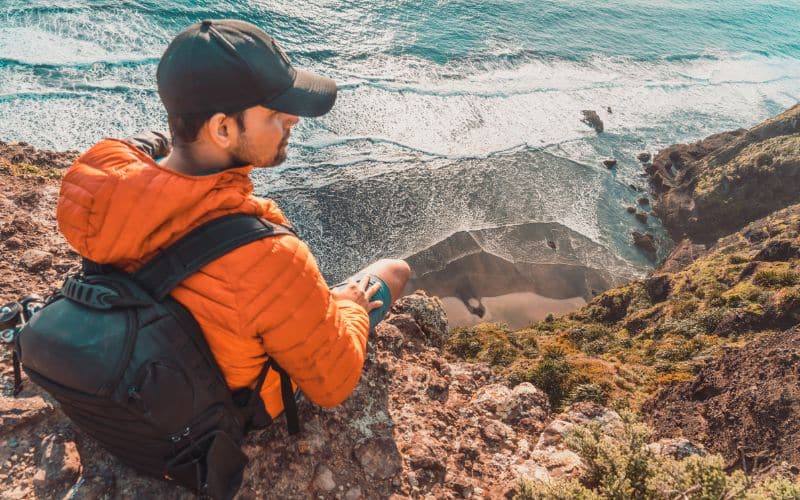 Qu'est-ce qu'un sac à dos ergonomique ? Les critères de choix Homme assit sur le haut d'une falaise, portant une casquette, une doudoune orange et un sac à dos foncé.