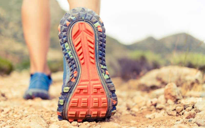 Photo de l'arrière d'un pied, portant une basket ergonomique, qui se soulève du sol, seule la pointe touche le sol. Sur un chemin de terre.