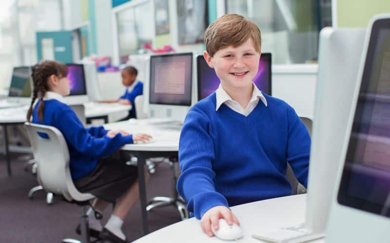 Jeune garçon, devant un ordinateur avec le sourire, en fond, on voit une jeune fille, elle aussi, devant un ordinateur.