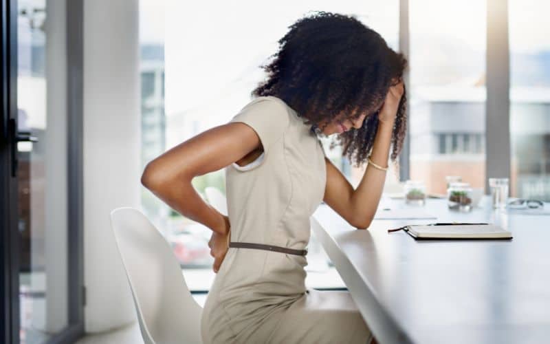 Femme assise à un bureau, elle se tient le dos et la tête. On voit qu'elle souffre.