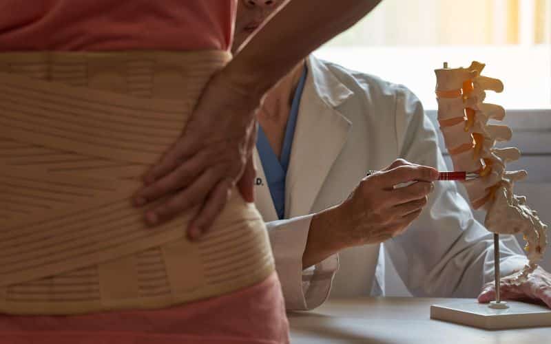Femme de dos portant une ceinture lombaires, face à un médecin qui lui montre un endroit spécifique de la colonne vertébrale sur une maquette.