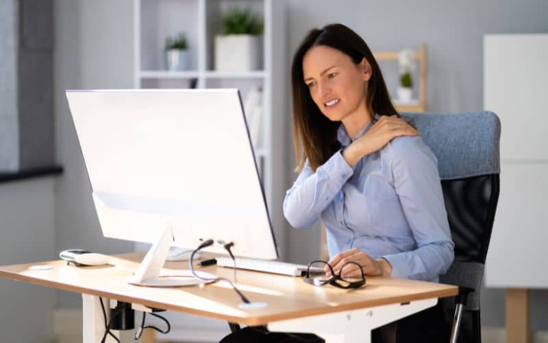 Femme derrière son bureau face à un ordinateur, elle travaille et elle fait une grimace en se touchant l'épaule.