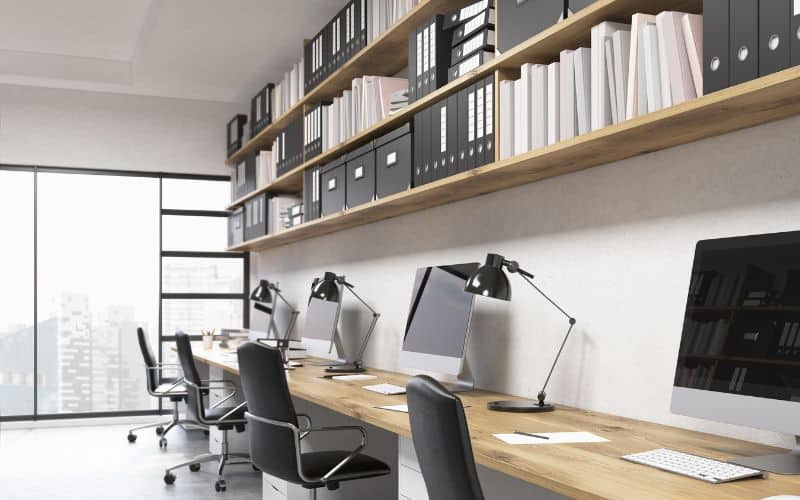 Bureau comun avec plusieurs ordinateurs et fauteuils ergonomiques, contre un mur,avec des étagères au dessus avec des dossiers bien rangés et organisés.