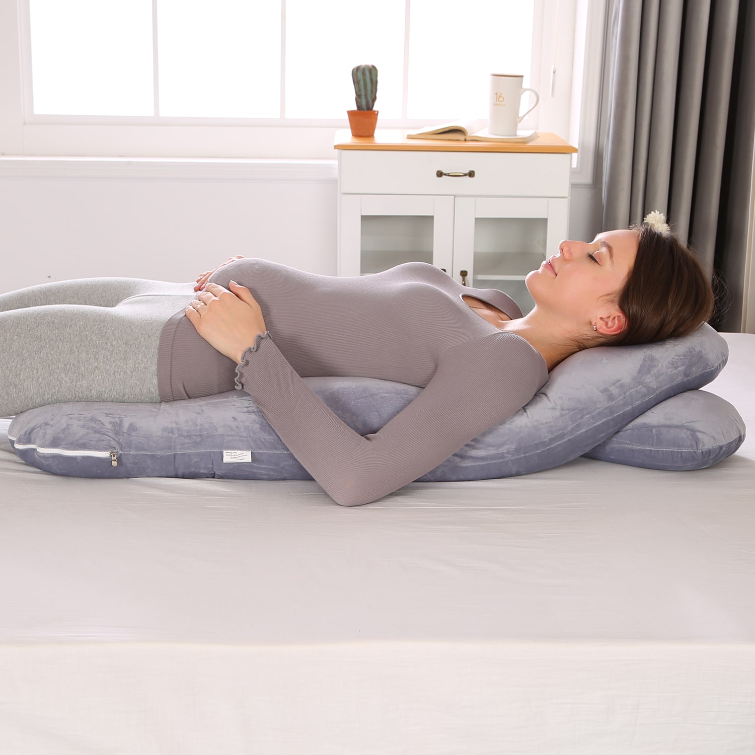 Comment bien positionner son coussin de grossesse pour dormir Femme enceinte allongée sur le côté avec un coussin de grossesse en U et un coussin plat sous le ventre, dans une chambre lumineuse.