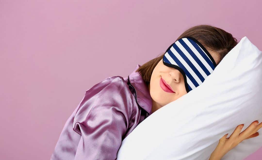 Comparatif oreiller ergonomique : le meilleur oreiller pour vous Femme endormie prenant sur elle un oreiller, yeux bandés à rayures, fond rose clair. Alt texte pour un comparatif oreiller ergonomique.